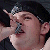Pixelated close-up of a person with dark hair wearing a dark top, mouth open in a shout or yell, hand raised near the face.