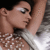 Pixelated close-up of a person with light skin tone, dark hair, and silver eyeshadow wearing a white top with brown patterned details.