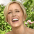 A pixelated close-up of a person with light brown hair smiling broadly, showing teeth, against a green foliage background with a pink flower on the right.