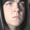 A static close-up of a person with light skin, dark hair, and light-colored eyes, wearing a dark hooded garment with visible light streaks on the left side.