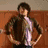 A pixelated image of a person with dark curly hair wearing a brown jacket over a light purple shirt, standing against a reddish-brown wall.