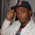 A person wearing a dark cap with a pink logo adjusts the cap with their right hand, wearing a white jacket over a colorful striped shirt against a blurred background.