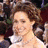 A woman in a red strapless gown smiles while posing on a red carpet with photographers visible behind her.