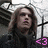 Pixelated static image of a person with long dark hair wearing dark clothing, standing before a microphone and a cross-shaped structure with a pink heart symbol.