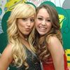 Two women with long hair pose closely together against a green and yellow patterned background.