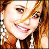 A close-up of a person with light brown hair, wearing large white dangling earrings and a white top, smiling with visible teeth.