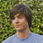 Pixelated static image of a person with short brown hair wearing a light blue shirt against a green foliage background.