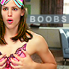 A person with long brown hair wears a colorful striped headband and a pink patterned top, hands near chest, in a room with a gray couch and curtains.