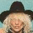Pixelated image of a person with light-colored hair wearing a dark wide-brimmed hat against a pale blue background.