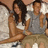 Two people sit side-by-side on a light-colored couch, the person on the left wearing a white top and dark pants, the person on the right in a gray sleeveless shirt and light shorts.