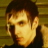 Pixelated close-up of a person with short dark hair, wearing a dark top against a yellowish-brown background.