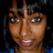 Pixelated close-up of a person with dark hair and brown skin, wearing blue clothing against a black background.