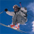 Pixelated static image of a skier in mid-air against blue background with red and white ski boots, black gloves, gray outfit, brown hair, and purple headband.