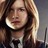 A pixelated close-up of a person with long brown hair, wearing a dark top with a light collar, against a blurred background.