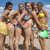 Group of five women in swimwear posing on a beach with blue sky and clouds, one woman holds green frisbee.