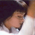A pixelated close-up of a person's face with light skin tone, dark hair, and a white garment, set against a dark background with light brown and white areas.