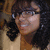 A person with dark curly hair and glasses smiles in a light-colored top against a blurred background.