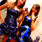 Two women in dark blue and blue outfits pose together, one holding a small object, with a light background.