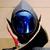 A stylized helmet with a blue reflective visor, gray top, and black base, featuring red accents on the sides, displayed in a