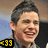 Pixelated close-up of a smiling person with short dark hair, wearing a dark jacket over a light shirt, with a yellow and black graphic containing '33' in the lower left corner.