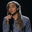 A pixelated image of a person with long brown hair wearing a light blue hoodie, holding a microphone against a dark background.
