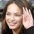 A pixelated close-up of a person with long brown hair smiling, holding a hand near their head against a dark background.
