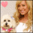 A woman with long blonde hair and a white top stands beside a small white dog wearing a pink bow tie, with a pink heart symbol in the background.