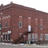 A pixelated static image of a red-brick building with multiple windows and a dark entrance, viewed from a low angle.