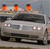 Static image of a silver sedan with three orange cylindrical objects on its roof against a blurred background.