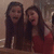 Two women in red dresses stand side-by-side, smiling with open mouths, against a blurred indoor background.