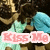 Two figures in close embrace with a pink banner displaying 'KISS ME' text, set against dark background.