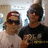 Two people pose together in a pixelated image with one person wearing sunglasses and a red cap, the other in dark clothing with visible text on the shirt.