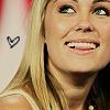 A close-up of a smiling person with long blonde hair, wearing a light-colored sleeveless top, with a small black heart symbol on the left side of the image.