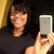 A person with dark hair and a black shirt holds a silver smartphone in front of their face, smiling at the camera against a yellow background.