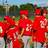 Group of people in red uniforms with white numbers on backs, standing outdoors under blue sky with trees and fence background.