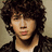 Pixelated portrait of a person with curly brown hair wearing a dark jacket over a light shirt against a beige background, with cyan text elements on a dark red backdrop.