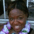 A pixelated close-up of a person with dark skin wearing a purple and white patterned shirt, set against an indistinct background.