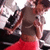 A person in a gray crop top and red skirt stands with arms crossed, head tilted down, against a blurred background of dark shapes.