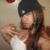 A person with long brown hair wearing a black beanie and white top, holding an object near their chest against a light background.