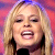Pixelated close-up of a person with long blonde hair holding a black microphone against a pink and purple gradient background.