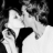 Black-and-white pixelated image of two figures in close physical contact with one figure's hand on the other's face, set against a dark background.