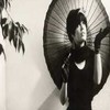 Black-and-white image of a person holding a large circular fan, with a leafy branch on the left side.