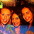 Three people in a group pose with smiling expressions, wearing blue and pink clothing against a dark background with a green logo element.