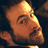 Pixelated close-up of a cartoon character's face with warm orange and brown tones, featuring visible facial features like an eye, eyebrow, nose, mouth, and beard against a dark background.