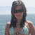 A woman in a light blue bikini top stands against an ocean backdrop, wearing dark sunglasses and long brown hair.