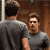 Two pixelated figures in gray shirts stand against a brown wooden background, one facing away and the other facing forward with a neutral expression.