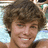 A pixelated image of a person with brown curly hair smiling, wearing a dark top against a light blue background with a pink symbol on the right side.