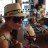 Pixelated static image of a person wearing a wide-brimmed hat and dark sunglasses, with a blurred background containing indistinct shapes and colors.