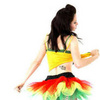 A woman with long dark hair wears a yellow crop top with green trim and a red-yellow-green tutu skirt, viewed from behind against a white background.
