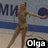 A female gymnast in a gold leotard performs with a blue ball on a light-colored floor, background shows partial text and blue stripes.